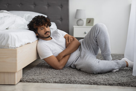 Pensive indian guy in pajamas sitting on floor by bed