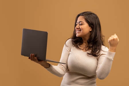 Overjoyed arab lady celebrating success with laptop, raising clenched fist, standing over beige backgroundの写真素材