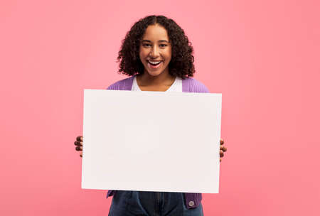 Happy millennial black woman holding empty poster, smiling at camera on pink studio background, mockupの写真素材