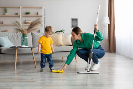 Little kid boy watching her mother mopping the floor after him. Woman wiping the floor with rag next to her babyの写真素材