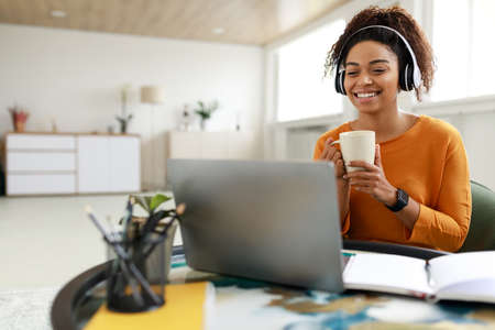 Smiling black lady watching video on computer, drinking coffee