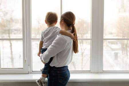 Young mother holding her little son on hands, standing near window and looking through it, back...