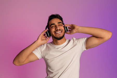 Peaceful young Arab guy listening to music in wireless headset, closing eyes and relaxing in neon lightの写真素材