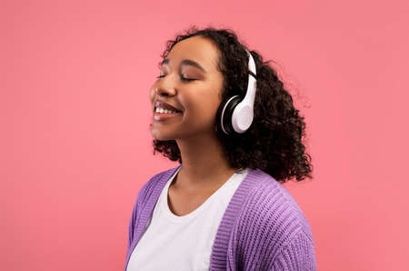 Lovely millennial black woman listening to music in wireless headphones, closing her eyes on pink studio backgroundの写真素材
