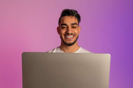 Handsome smiling young Arab man working on laptop, having video call, chatting online in neon lightの写真素材