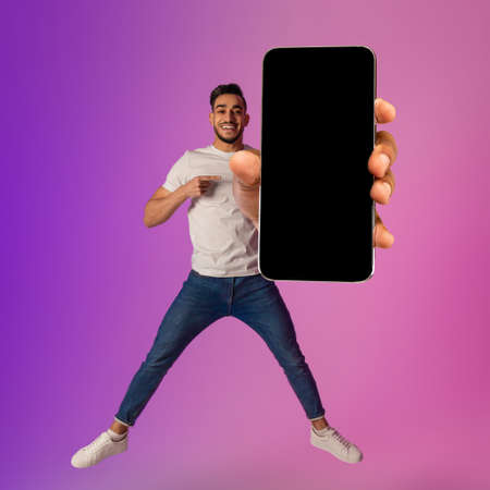 Handsome Arab guy showing big cellphone with empty black screen, jumping in air, offering space for mockup in neon lightの写真素材