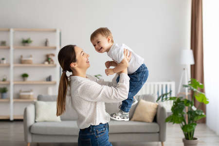 Emotional young caucasian mother raising laughing toddler son, having fun and playing together in...