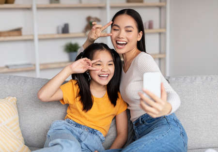 Satisfied teenage girl and young asian female have fun together, make selfie on smartphoneの写真素材