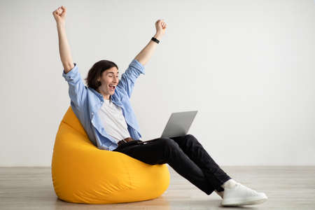 Excited man making YES gesture, sitting in beanbag chair with laptop, celebrating success or achievement, copy spaceの写真素材
