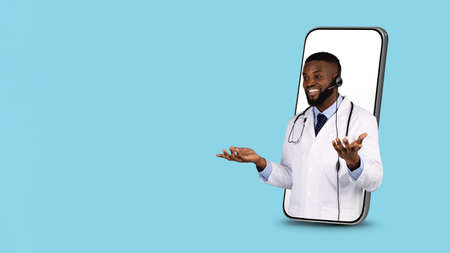 Telemedicine Concept. Black Doctor In Headset Peeking Out Of Big Smartphone Screenの写真素材