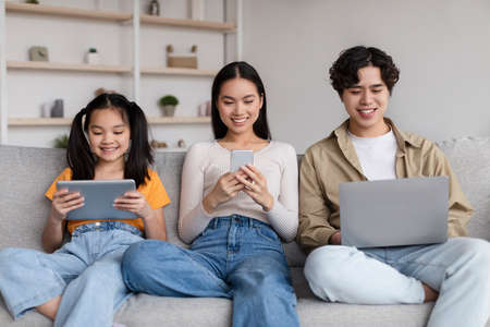 Smiling teen child and young asian mother and father surfing in internet, chatting on gadgets, play...