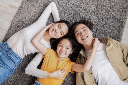 Satisfied millennial japanese mother, father and teen daughter lie on floor on gray carpet in room interiorの写真素材