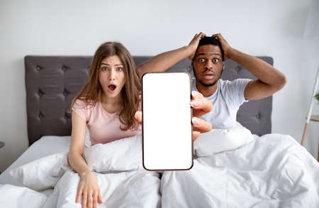 Shocked mixed race couple sitting in bed with smartphone, mockup