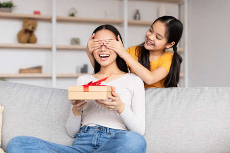 Smiling teen girl congratulates surprised young asian lady, gives box with present, closes eyes to momの写真素材