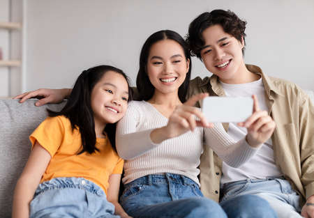 Glad smiling young asian couple and teen daughter making photo on phone on sofa in living roomの写真素材
