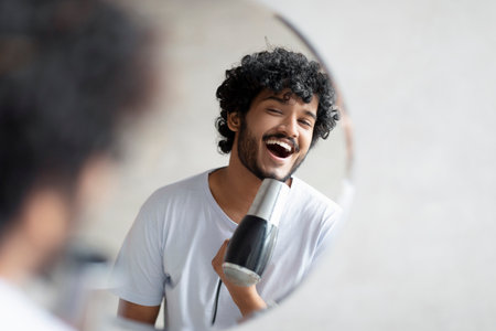 Joyful indian man holding hairdryer and singing as in microphone, having fun while doing morning beauty routineの写真素材