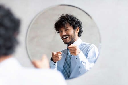 Youre handsome. Confident indian businessman pointing at mirror in bathroom with two fingers, wearing shirt and smilingの写真素材