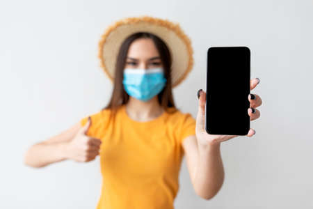 Young woman wearing protective face mask and hat, showing smartphone with empty blank screen on light wallの写真素材