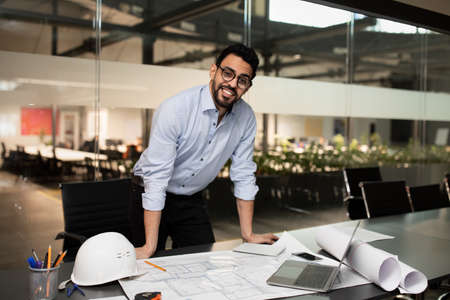Smiling millennial attractive muslim engineer builder in glasses working on desk with project drawingsの写真素材