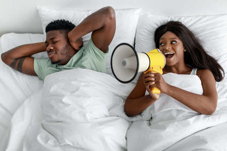 Sad young african american guy closes his ears on white bed, cheerful woman shouts at megaphone in bedroomの写真素材