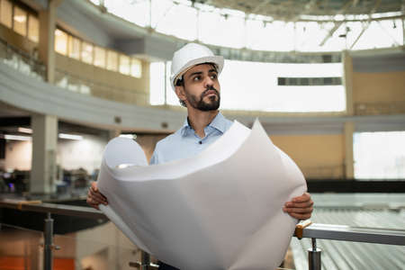 Serious busy young attractive arabic industrial engineer in hard hat with project drawings in factory interiorの写真素材