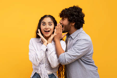 Shock, gossip, share advice. Young indian man whispering to lady on ear, surprised woman with open mouthの写真素材