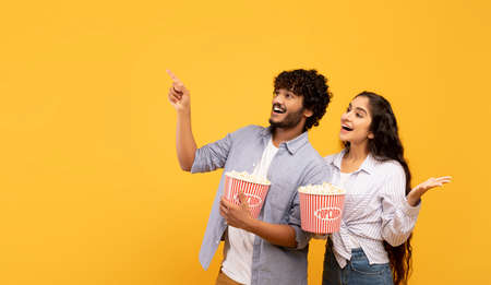 Happy indian couple eating popcorn, cheerful guy pointing at free space for advert or text, panoramaの写真素材