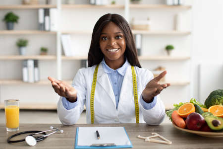 Healthy nutrition concept. Friendly black nutritionist giving online weight loss consultations, sitting at workplaceの写真素材