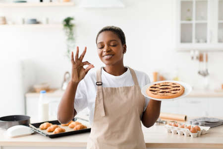 Satisfied millennial pretty black lady in apron enjoys aroma of fresh tasty pie in modern kitchen interior, free spaceの写真素材