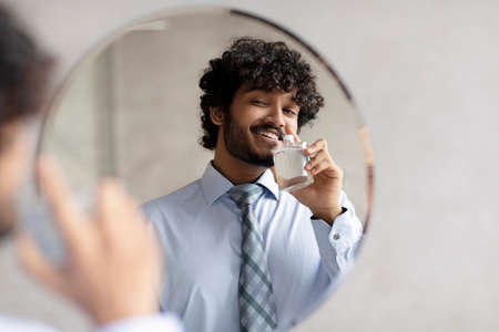 Handsome indian businessman in classic shirt and necktie smelling perfume while looking in the mirror in bathroomの写真素材