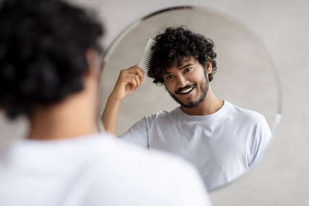 Male haircare. Positive indian bearded guy brushing hair with hairbrush, looking at his reflection in mirror in bathroomの写真素材