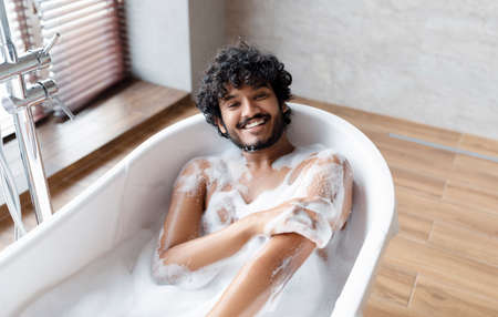 Happy naked indian man taking bath at home, laying in white bathtub with foam and smiling at camera, copy spaceの写真素材