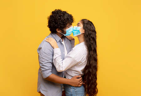 Love during pandemic. Young indian couple kissing and hugging, wearing medical face masks over yellow backgroundの写真素材