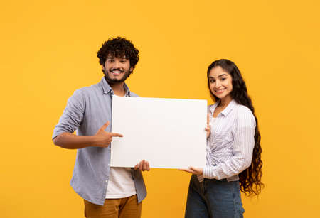 Place for your ad. Portrait of indian couple holding empty blank placard board, pointing at it over yellow backgroundの写真素材