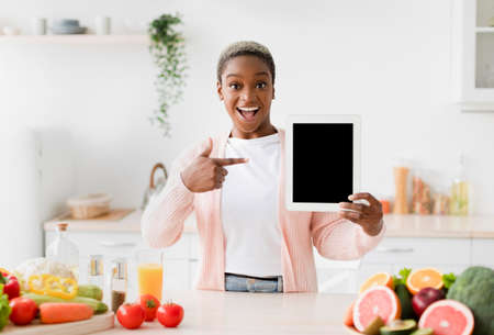 Glad excited emotional young pretty black woman points finger at tablet with blank screen at kitchen interiorの写真素材