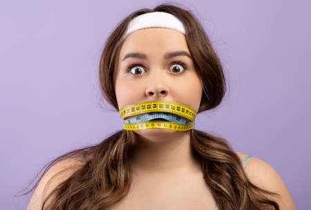 Shocked frightened pretty hungry caucasian plus size young woman with closed mouth with measuring tapeの写真素材
