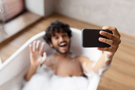 Joyful indian man lying in bathtub and waving hand on smartphone webcamera, resting in bath filled with water and foamの写真素材