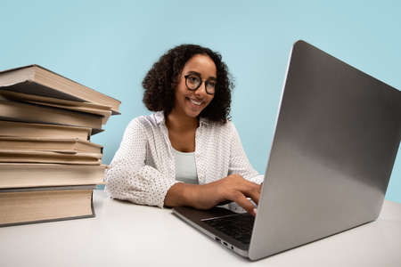 Positive young black female student with heap of books working on laptop, preparing for online exam...