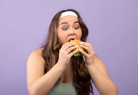 Funny hungry caucasian plus size young female in sports uniform greedily eats burgerの写真素材