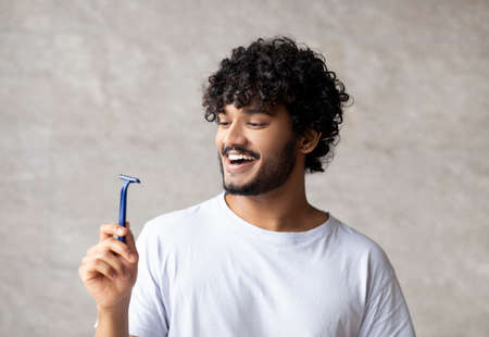 Body care concept. Happy indian man holding razor and smiling, standing in bathroom interior, copy...