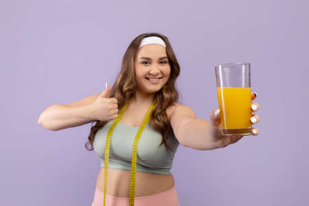Smiling european plus size young female in sportswear with measuring tape show glass of fresh orange...