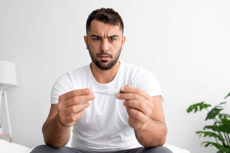 Sad shocked young male with beard looks at thermometer with high temperature and suffering from ill...