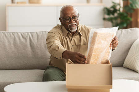Mature African American Male Unpacking Box Sitting On Sofa Indoorの写真素材