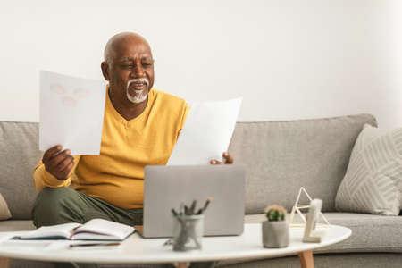 Senior Black Male Holding Two Papers Working On Laptop Indoor