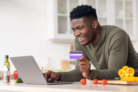Ordering Food Online. Black Guy Using Laptop And Credit Card In Kitchen