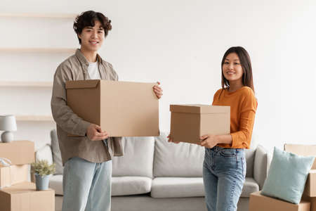 Cheerful young Asian woman and her husband holding carton boxes, smiling at camera in new home....