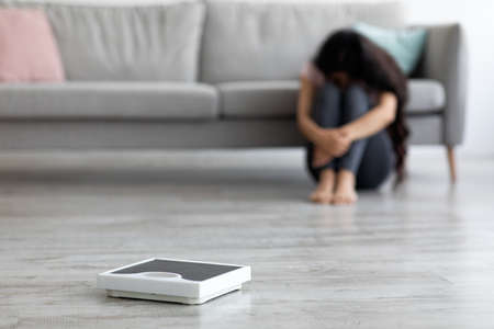 Unhappy young Indian woman crying, failing to lose weight, sitting on floor at home, selective focus on scalesの写真素材