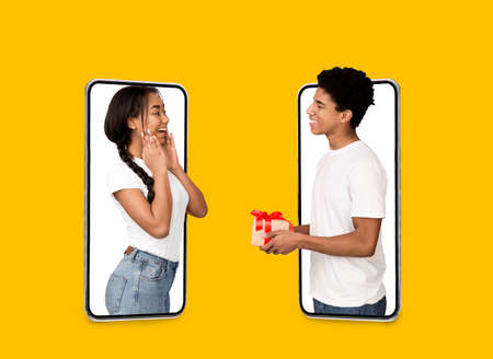 Smiling young black guy gives gift to surprised woman on blank screens of large phone isolated on...