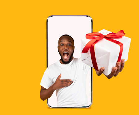Millennial funny african american male with open mouth gives gift in box and shouts empty screen of big phoneの写真素材