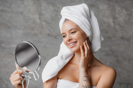 Beauty rituals. Happy lady with bath towel on head touching her glowing skin and looking in magnifying mirrorの写真素材